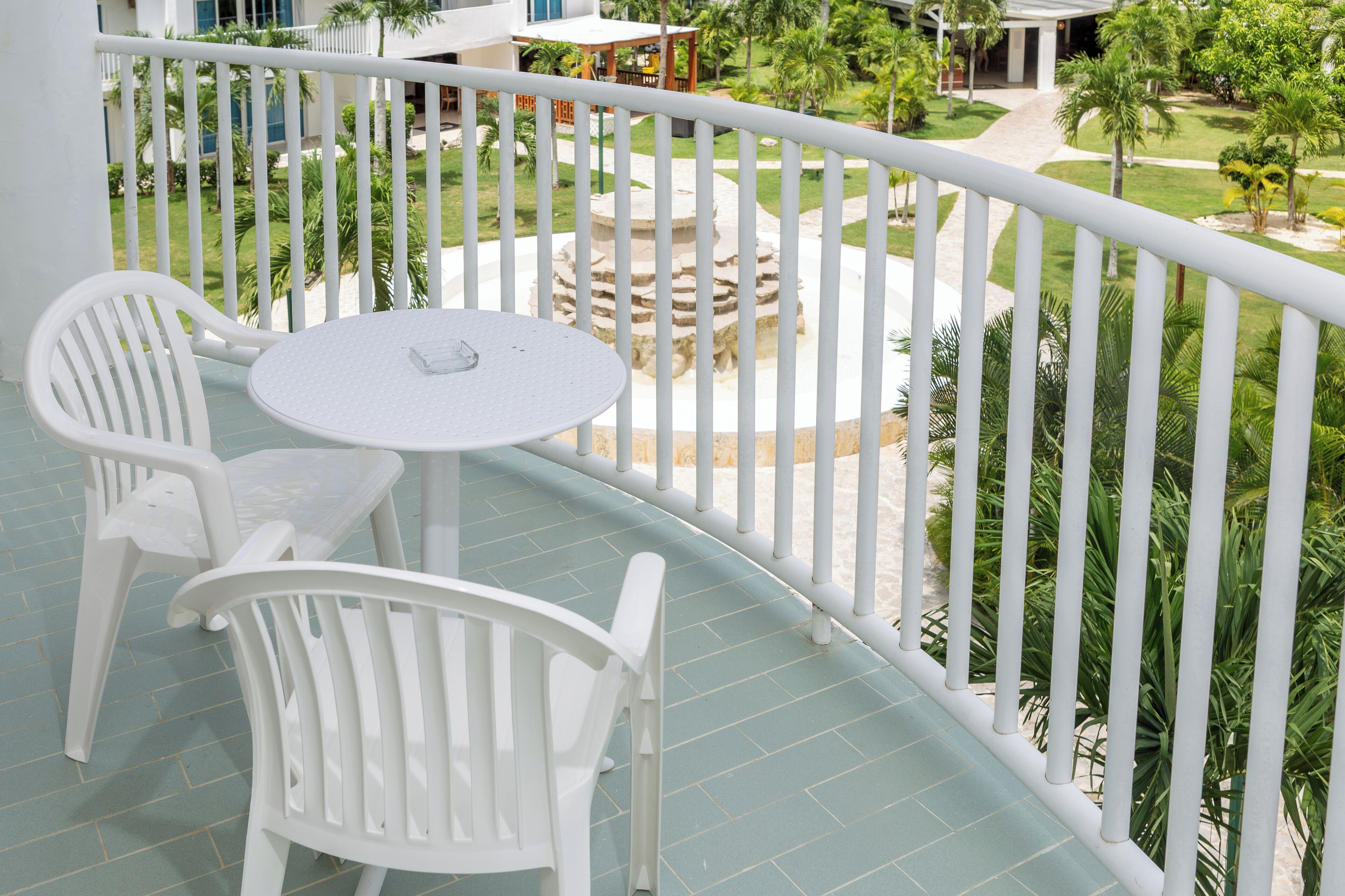 Uma varanda com cadeiras de plástico branco e uma pequena mesa redonda, com vista para um jardim arborizado com palmeiras e uma fonte de pedra.