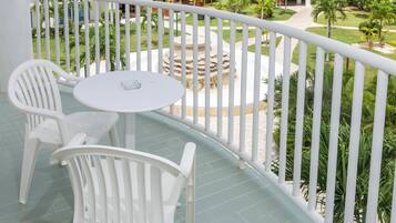 Uma varanda com cadeiras de plástico branco e uma pequena mesa redonda, com vista para um jardim arborizado com palmeiras e uma fonte de pedra.