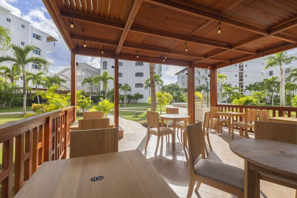 An outdoor patio area with wooden tables and chairs, a wooden pergola, and a view of a residential building complex with palm trees. - HM Alma de Bayahibe - Adults Only - All Inclusive (San Rafael del Yuma)