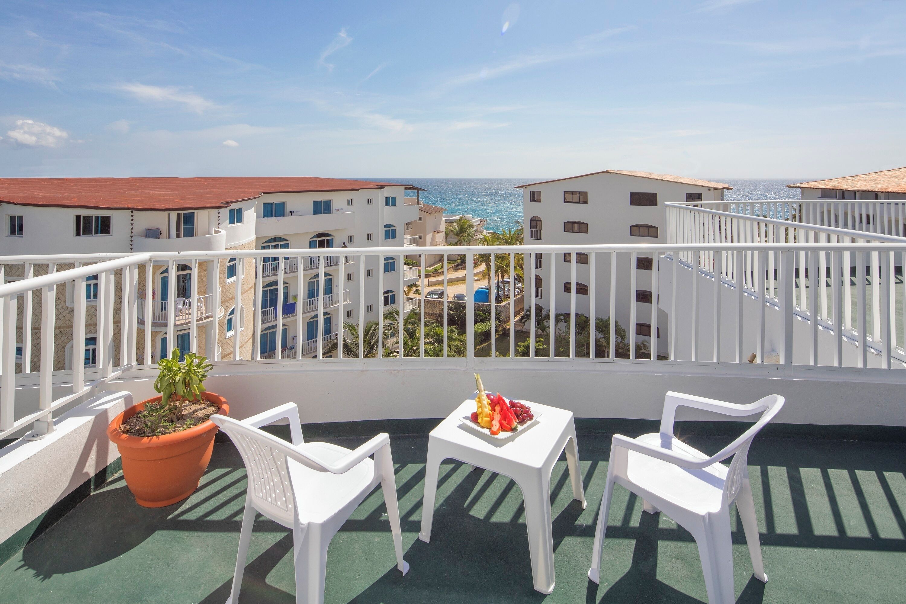 A balcony with two white plastic chairs, a small table with fruit and a potted plant, and a view of the sea and buildings.