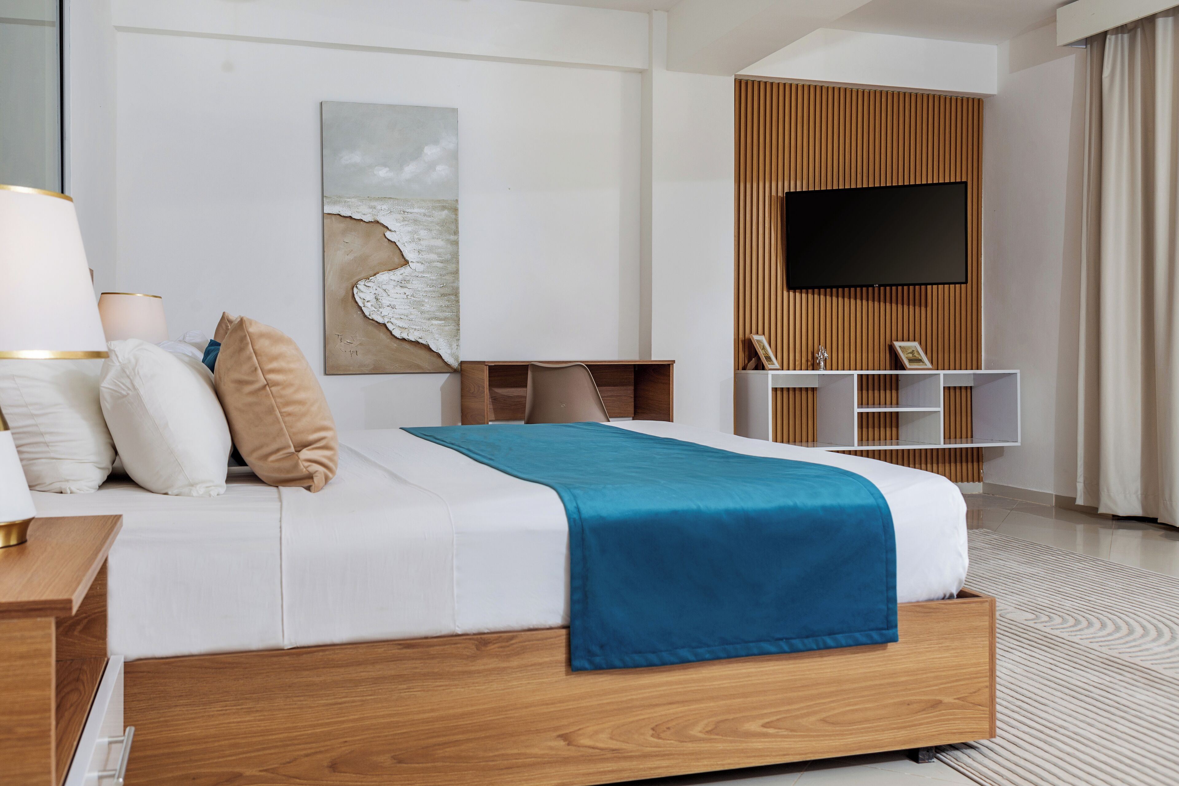 A modern hotel room with a large bed, a flat-screen TV, a wooden panel wall, and a beach-themed artwork.