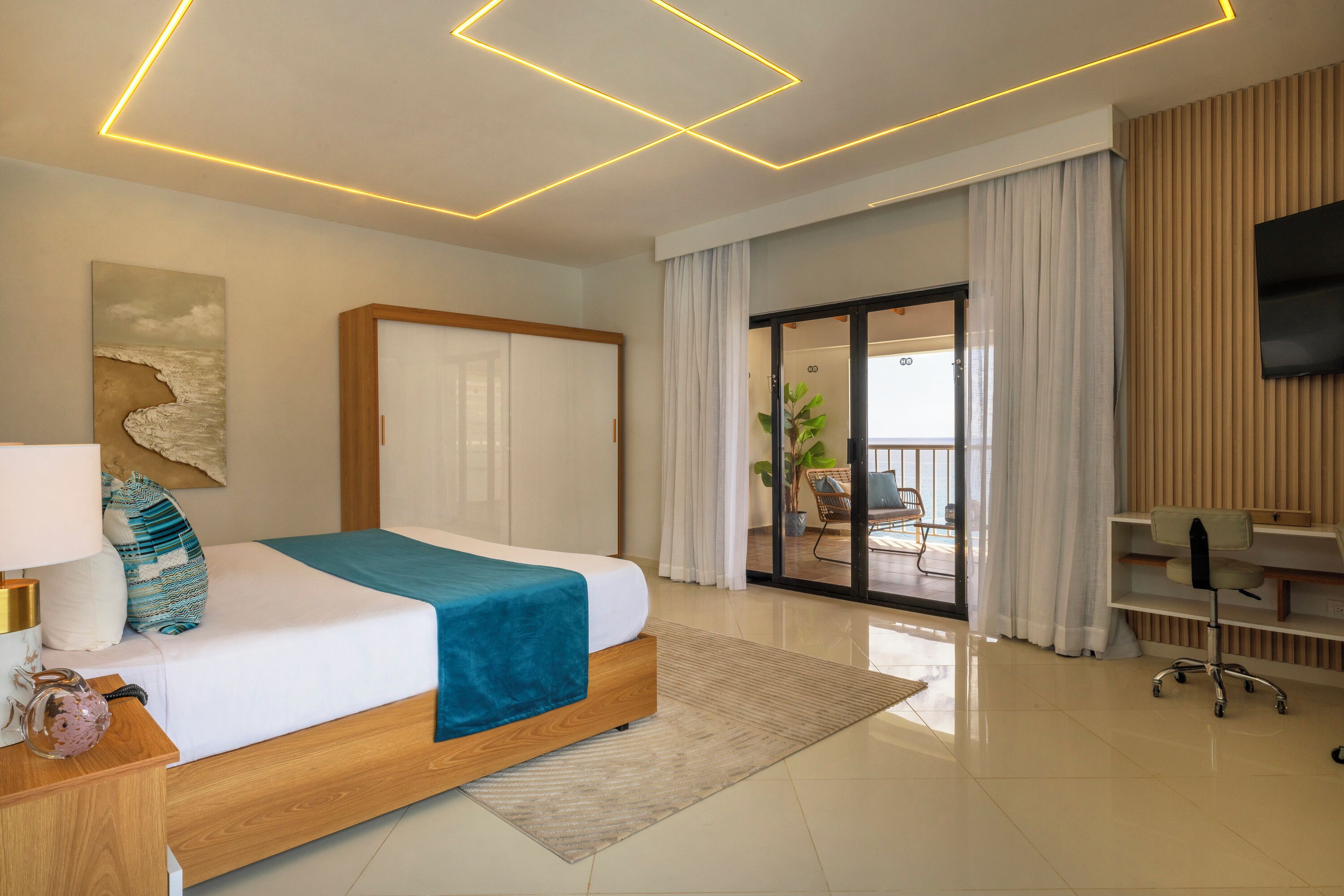 A modern hotel room with a bed, a desk, a chair, a TV, and a sliding glass door to the balcony.