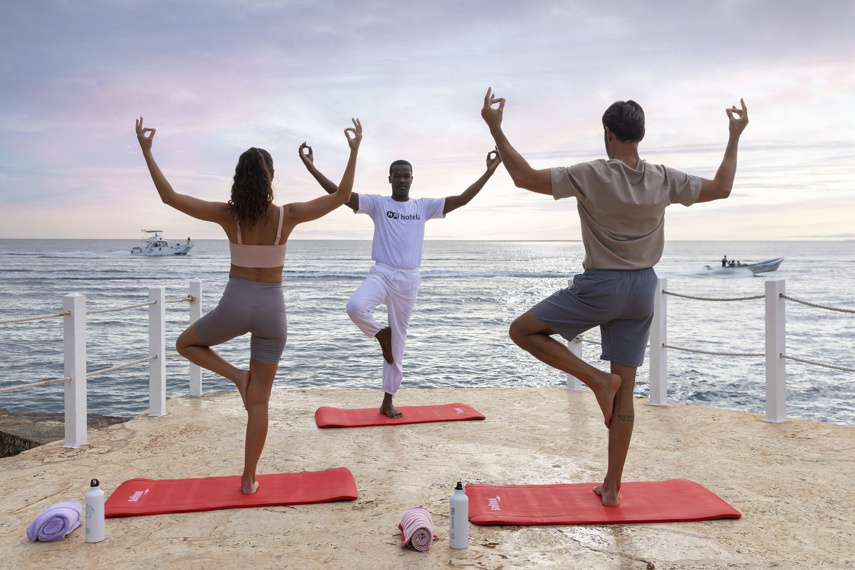 Three people performing yoga on red mats at a beachside location with boats in the background.