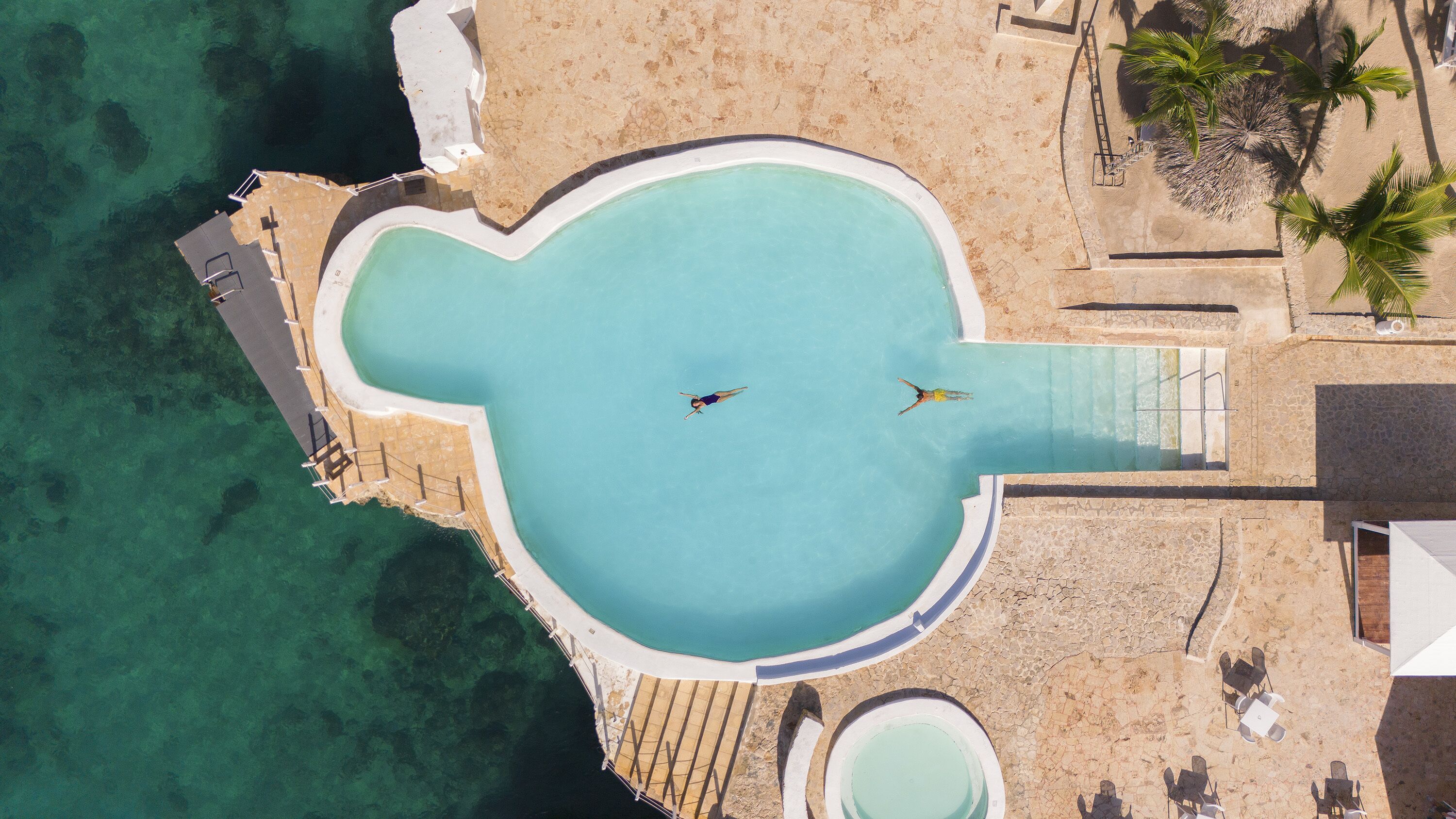 Eine Luftaufnahme eines Resorts mit einem Pool in ungewöhnlicher Form, einem Strandbereich und einem kleinen Bootssteg.