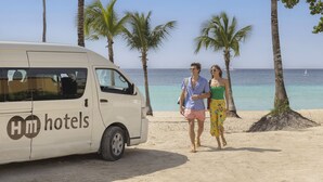 A white van with "HOTELS" branding parked on a sandy beach with palm trees and the ocean in the background. - HM Alma de Bayahibe - Adults Only - All Inclusive (San Rafael del Yuma)