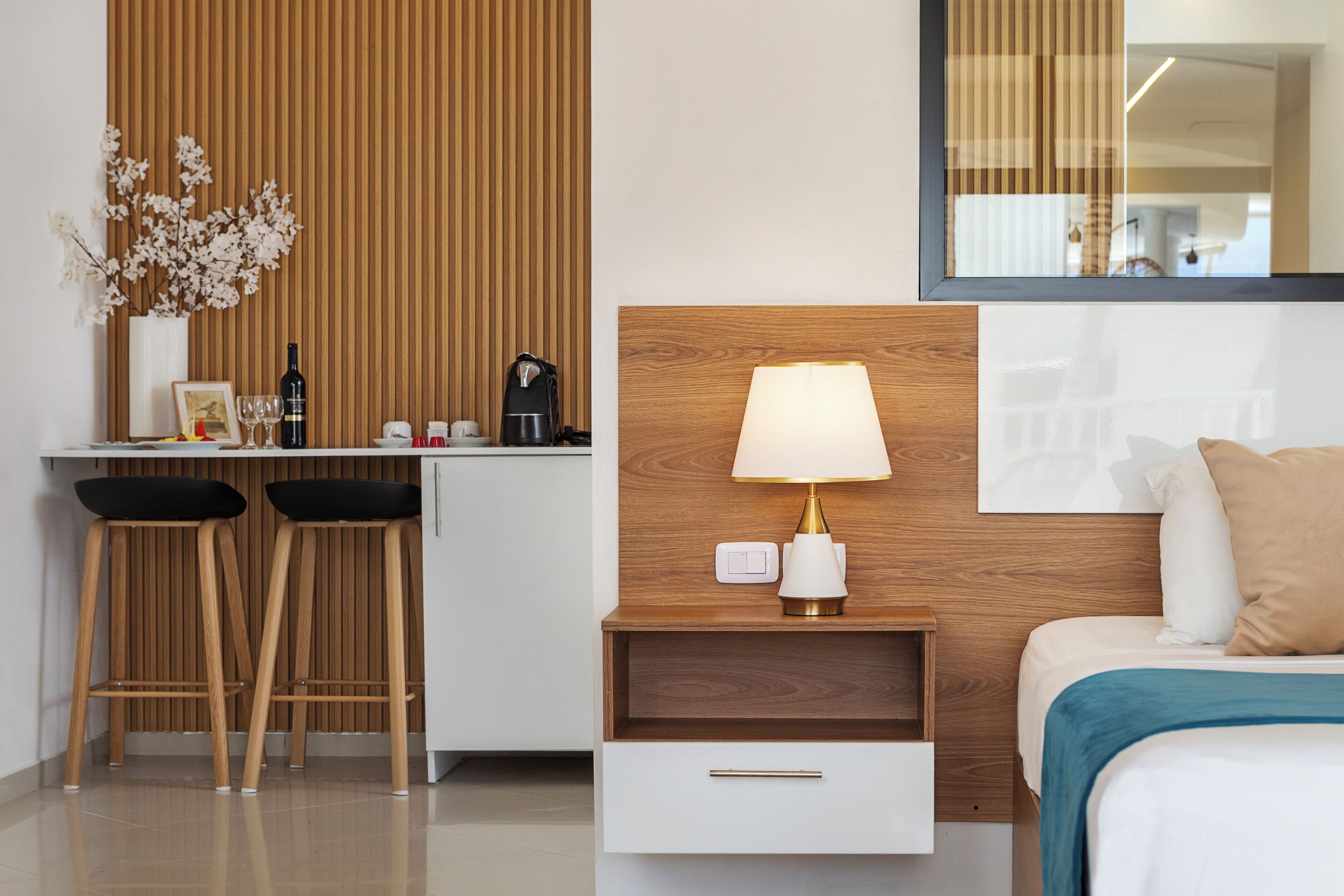 Une chambre d’hôtel moderne avec une tête de lit en bois, une table de chevet avec une lampe, un petit tiroir et deux tabourets de bar noirs.