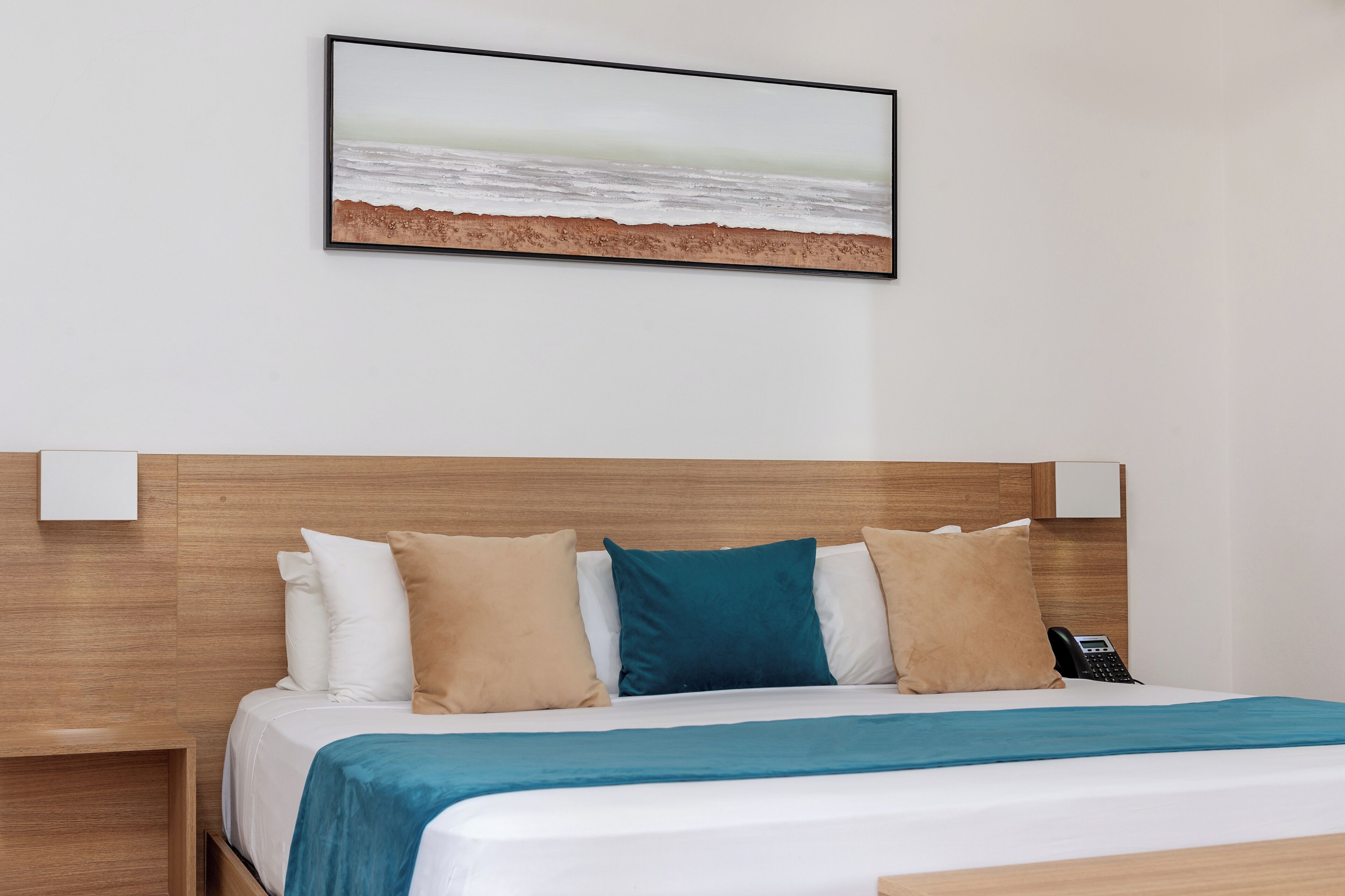 Habitació d'hotel amb un capçal de llit de fusta, un llit amb roba de llit blanca i aquamarina, i un quadre amb una escena de platja sobre el llit.