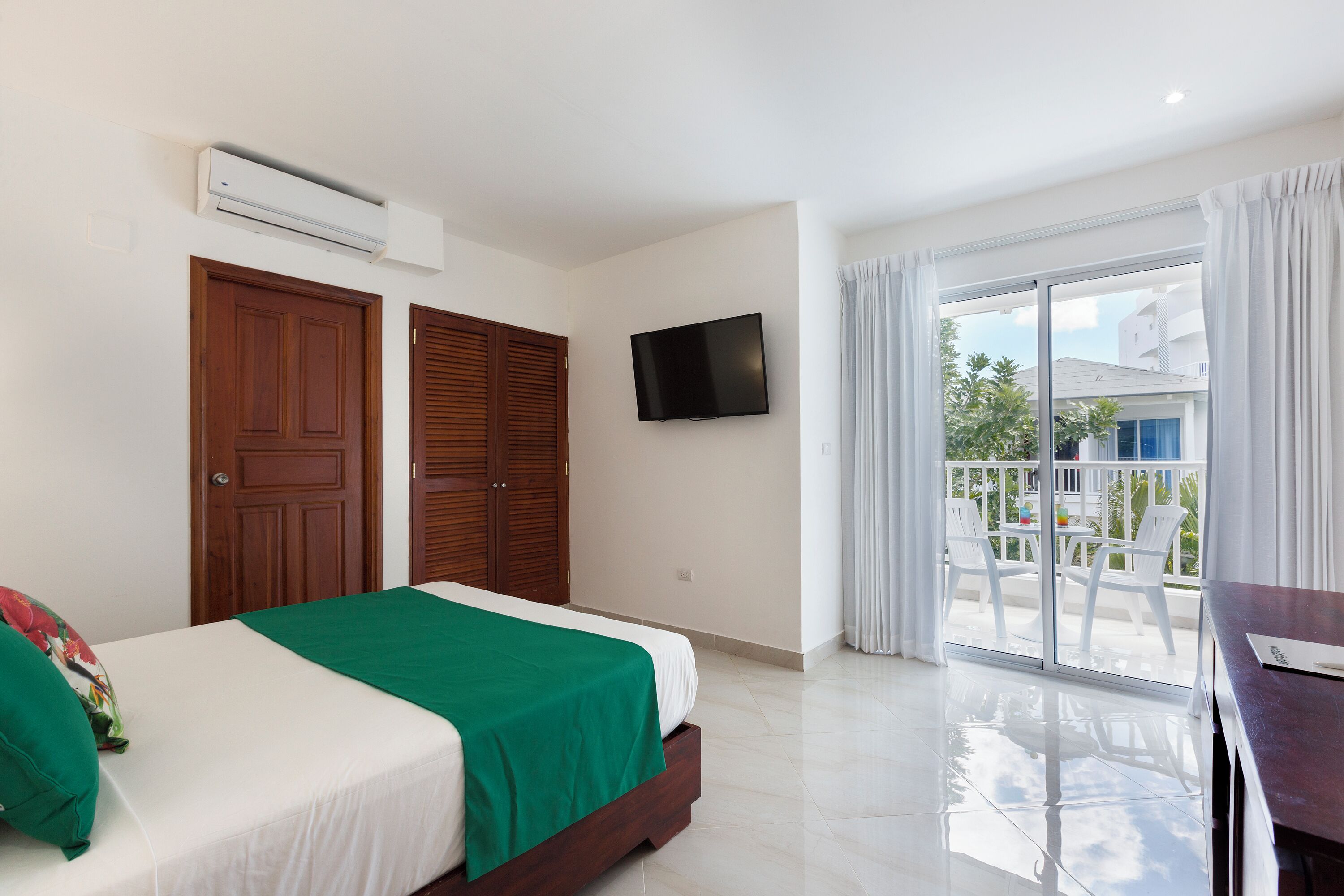 A hotel room with a bed, a TV, a balcony with chairs, and a sliding glass door.