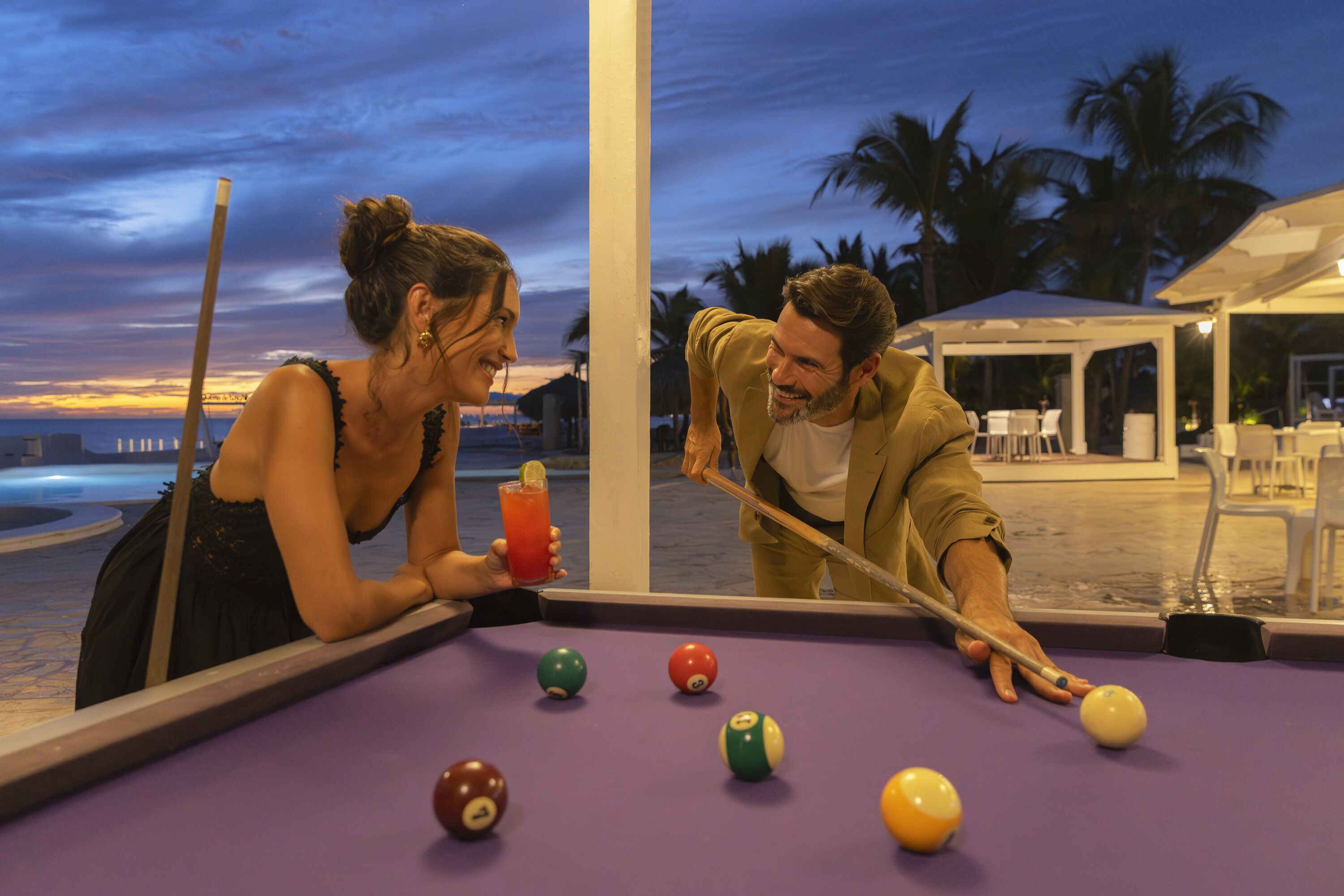 A pool table with balls, a drink, and a person leaning over the table.
