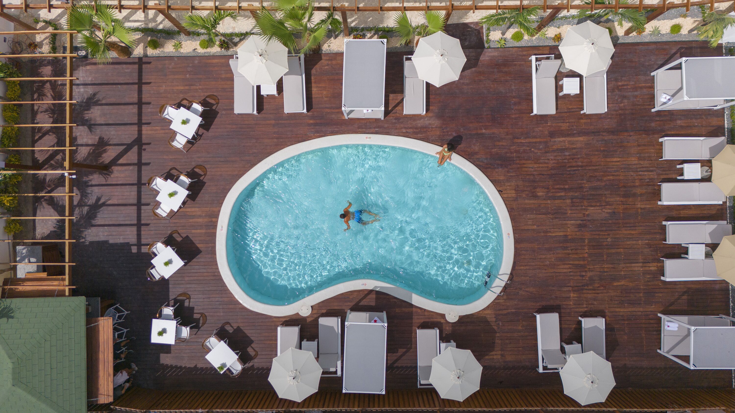 Vue aérienne d’un espace piscine avec une terrasse en bois, des chaises longues, des parasols et une petite piscine.