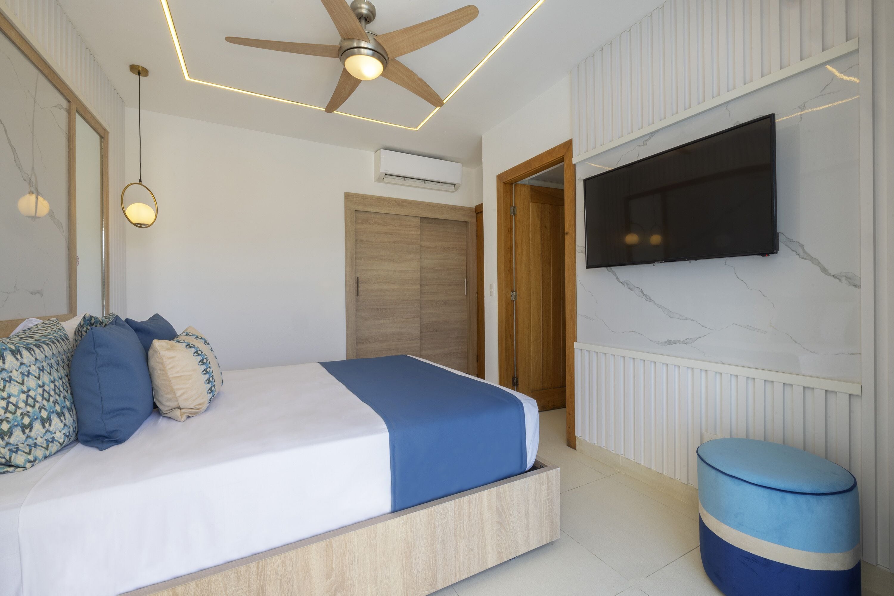 A bedroom with a bed, a ceiling fan, a television, and a small round ottoman.