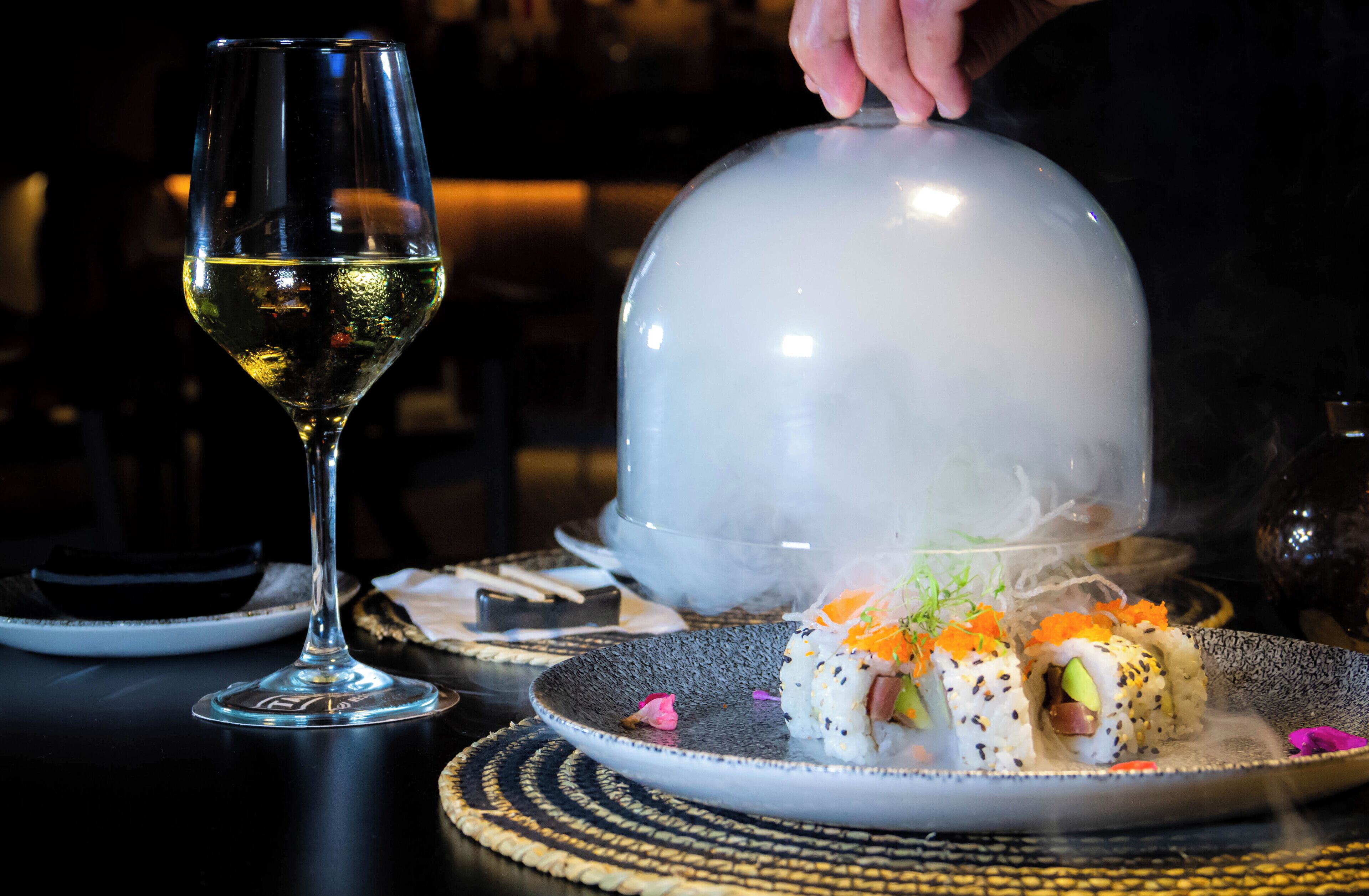 A dining table with sushi, a glass of white wine, and a decorative dome.