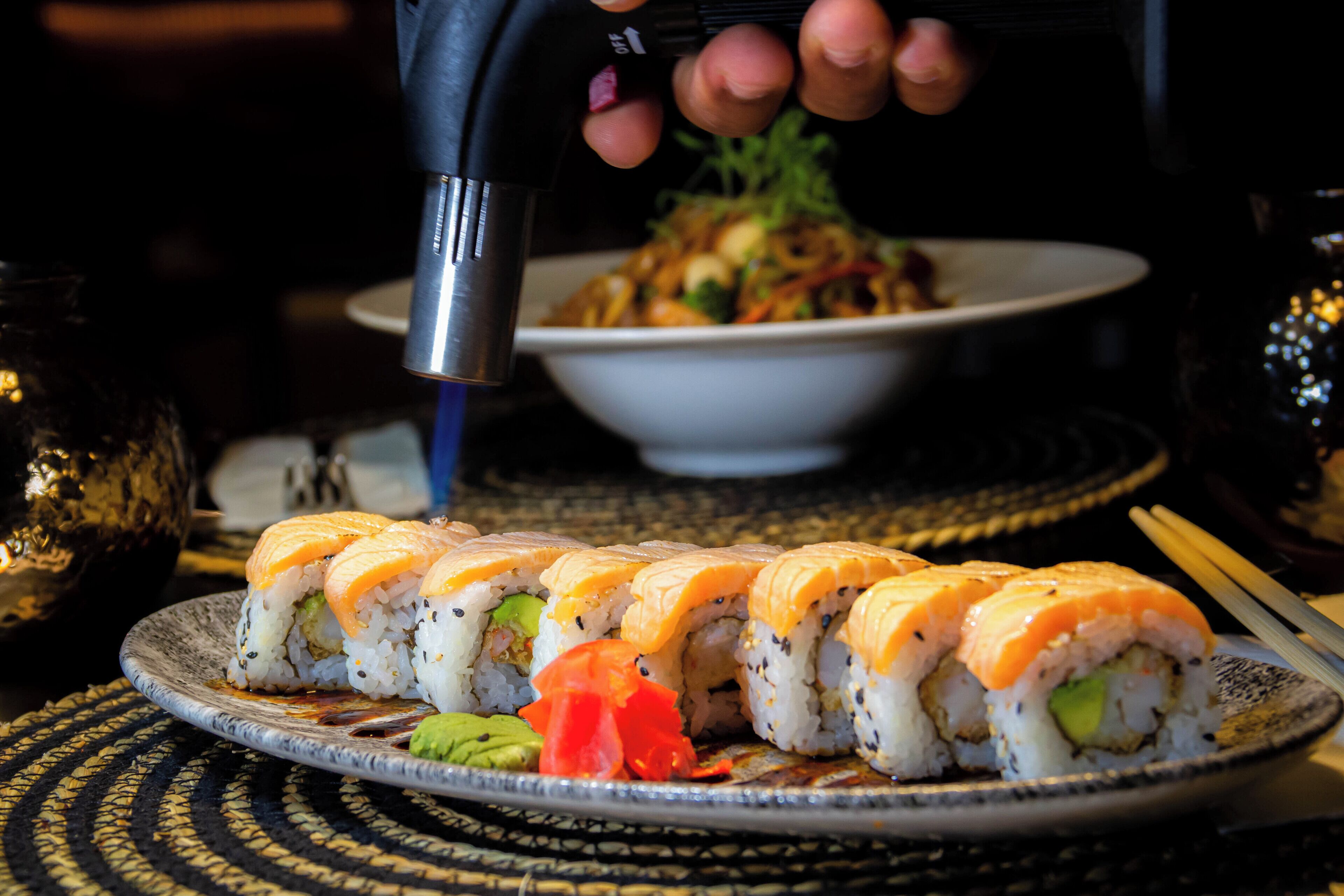 Een bord sushirollen met avocado en zalm, een kom roerbakgroenten en stokjes op een geweven placemat.