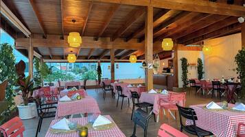 Un restaurante al aire libre con mesas a cuadros rojas y blancas, vigas de madera, luces colgantes y plantas en macetas.
