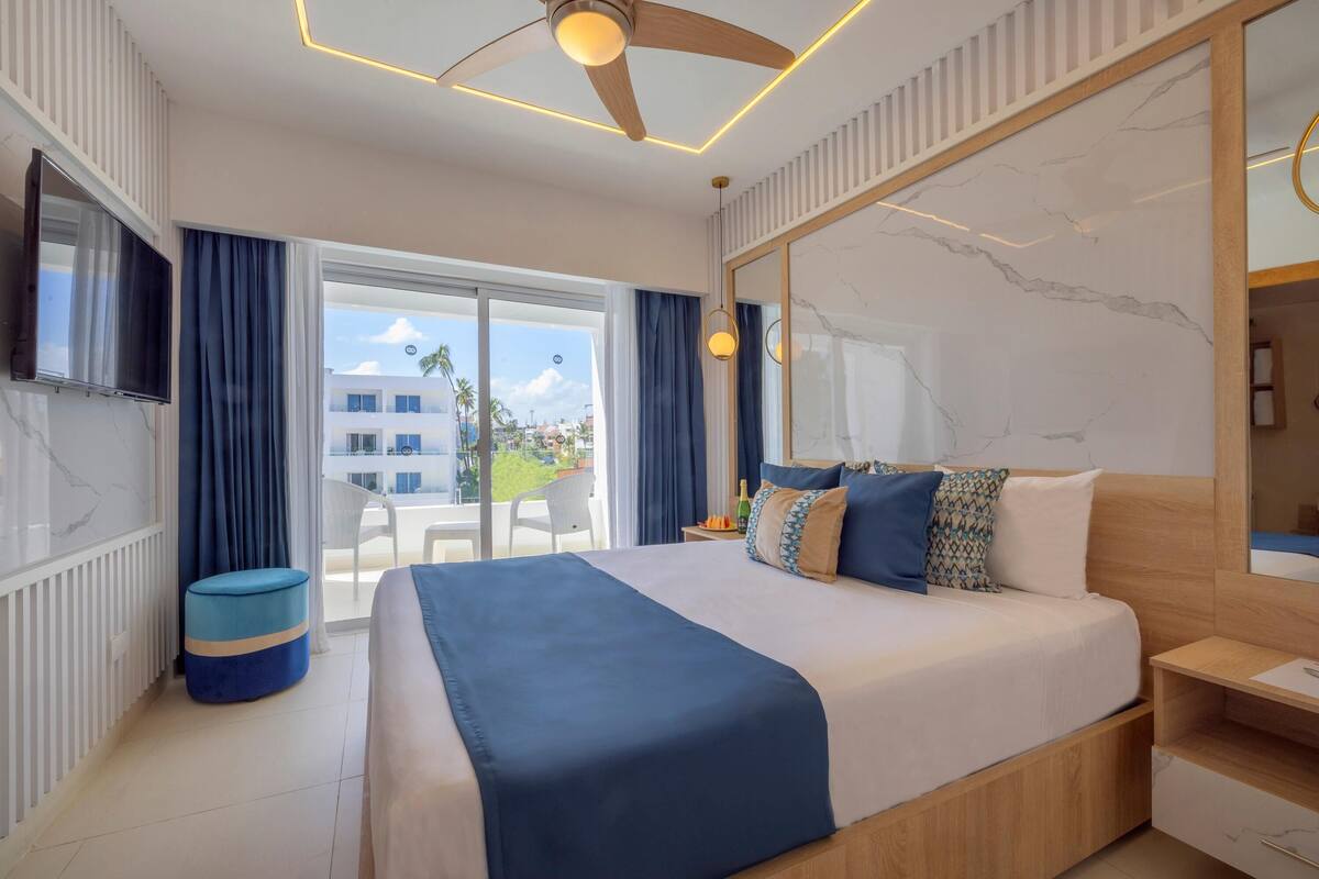 Habitación de hotel con una cama grande, ventilador de techo, televisor, ventana con cortinas y vistas al exterior.