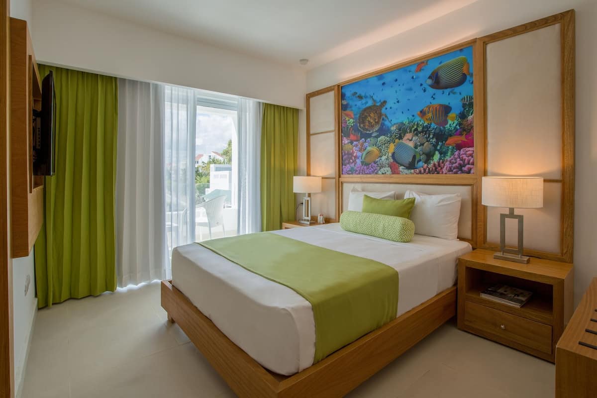 Una habitación de hotel con cama, dos mesitas de noche, televisión, un cuadro con escena submarina y ventana con cortinas.