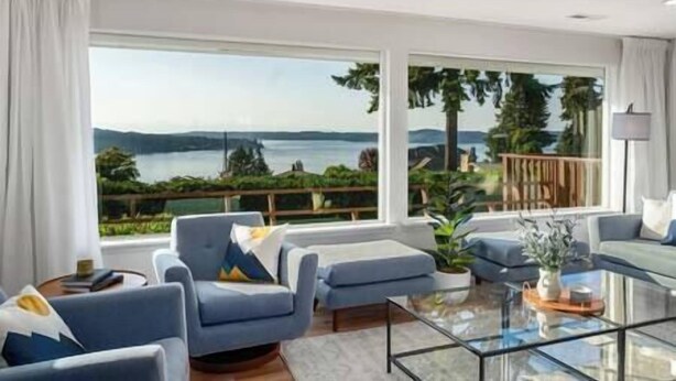 A modern living room with a large window offering a scenic view of a lake and mountains. - 5BR/4BA: Spacious Step-Free Main Level, Relaxing Views, Next to Chamber Bay Golf (University Place)