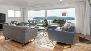 A modern living room with a sofa, armchairs, and a coffee table. There is a large window with a view of the sea. - 5BR/4BA: Spacious Step-Free Main Level, Relaxing Views, Next to Chamber Bay Golf (University Place)
