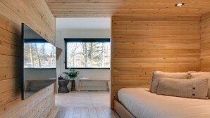 Ein Interieur einer Holzhütte mit einem Bett, einer Bank, einem Spiegel und einem Fenster mit Blick auf Bäume.