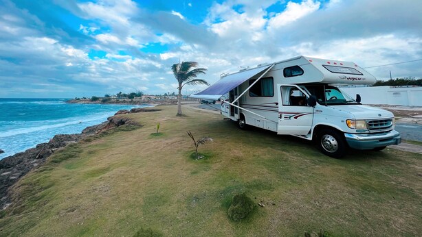 A white RV parked on a grassy area near the ocean with a clear sky. - Travel PR! (San Juan)