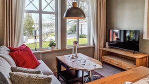 Una sala de estar con un sofá, una mesa con un candelabro, un televisor sobre un mueble de madera y ventanas amplias con cortinas.