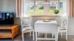 Un comedor con mesa y sillas, televisor y vistas a un jardín a través de amplias ventanas.