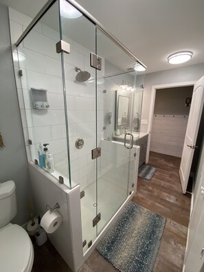 Ein modernes Badezimmer mit einer in Glas abgeschlossenen Dusche, weißen Fliesenwänden, Holzboden und einem sichtbaren Toilettenpapierhalter.
