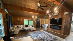 A spacious living room with a large sectional sofa, a coffee table, and a patterned rug. There is a ceiling fan, a TV mounted on the wall, and a door leading to another room. - Shore Thing Waterfront Home with private covered boat dock on Toledo Bend (Hemphill)