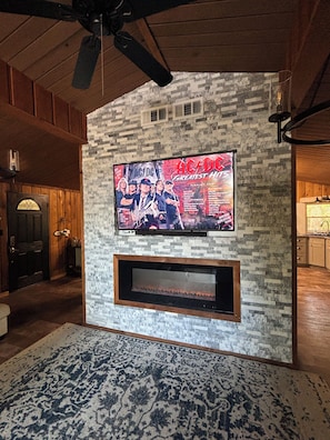 A living room with a stone wall, a flat-screen TV mounted above a fireplace, and a ceiling fan. - Shore Thing Waterfront Home with private covered boat dock on Toledo Bend Cove (Hemphill)