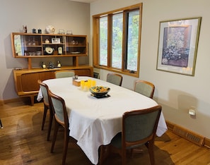 Een eetkamer met een ronde tafel, stoelen, een houten buffetkast en een raam met uitzicht op bomen.