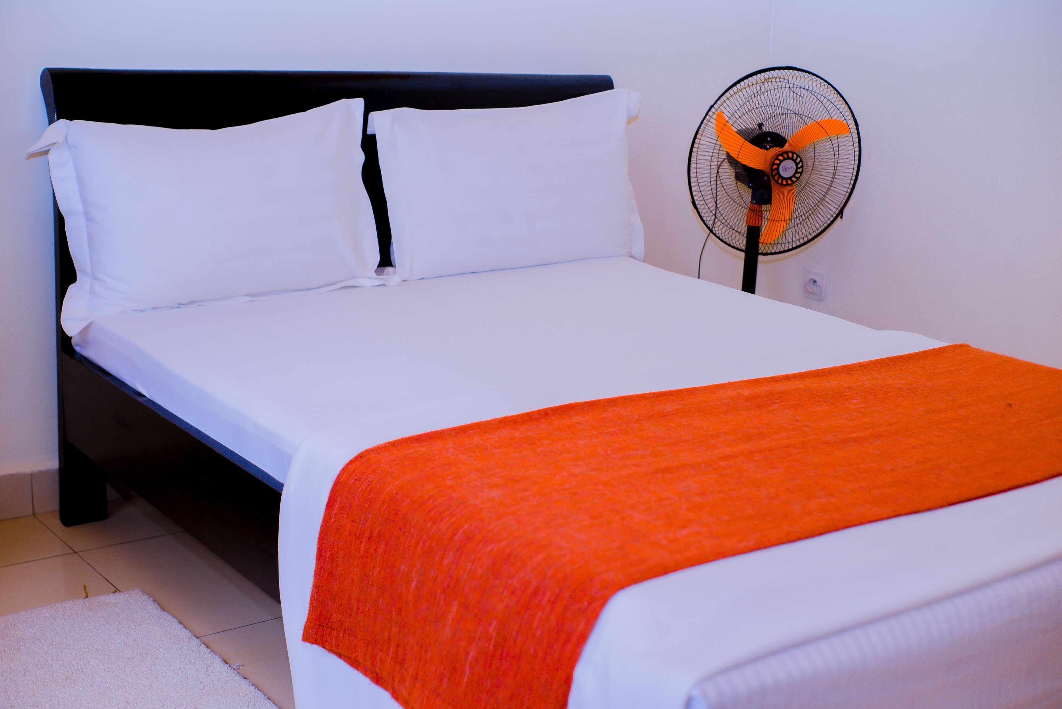A bed with white bedding and an orange throw blanket, a black headboard, and a standing fan with an orange fan blade.