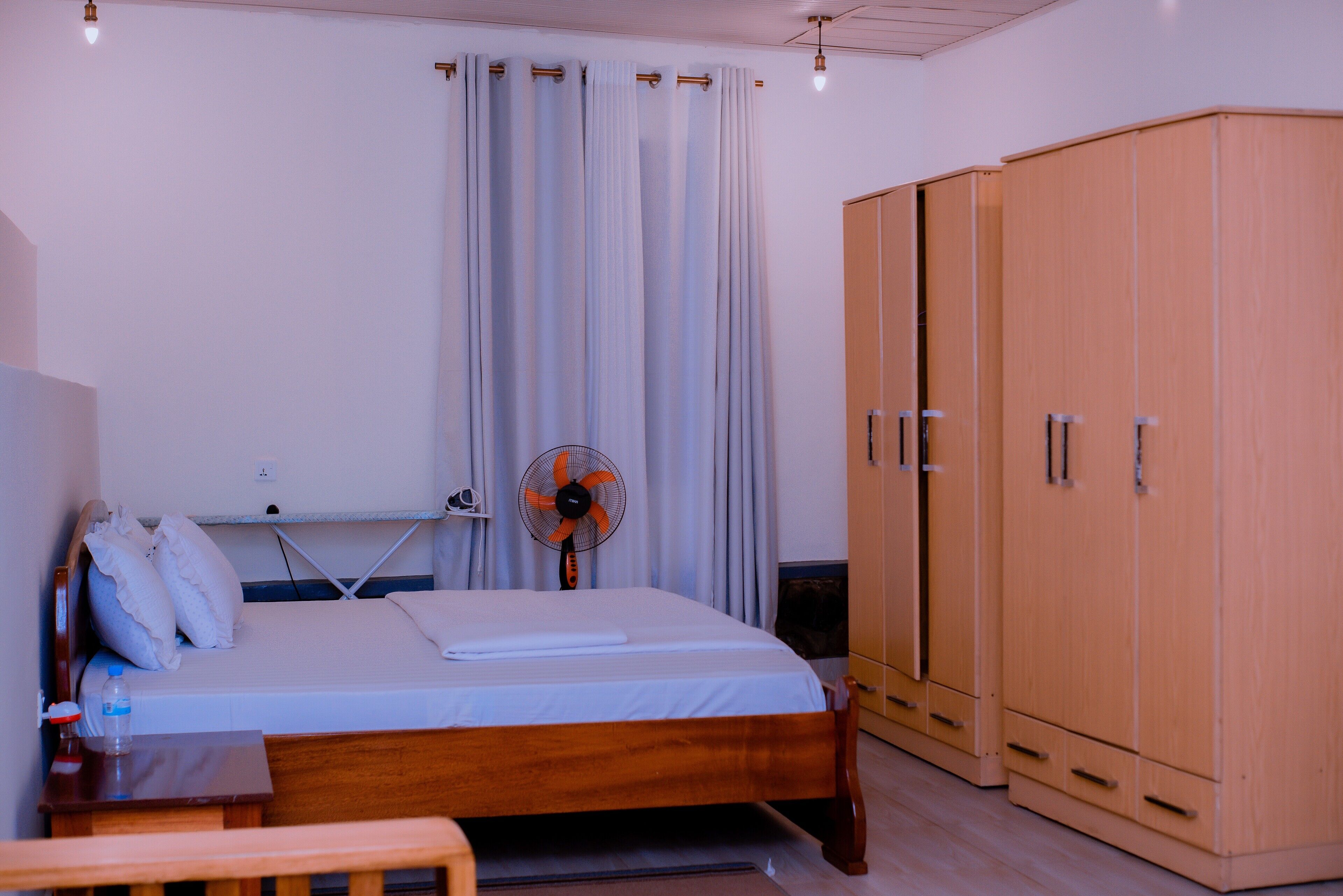 A bedroom with a bed, a wooden wardrobe, a fan, and a table.