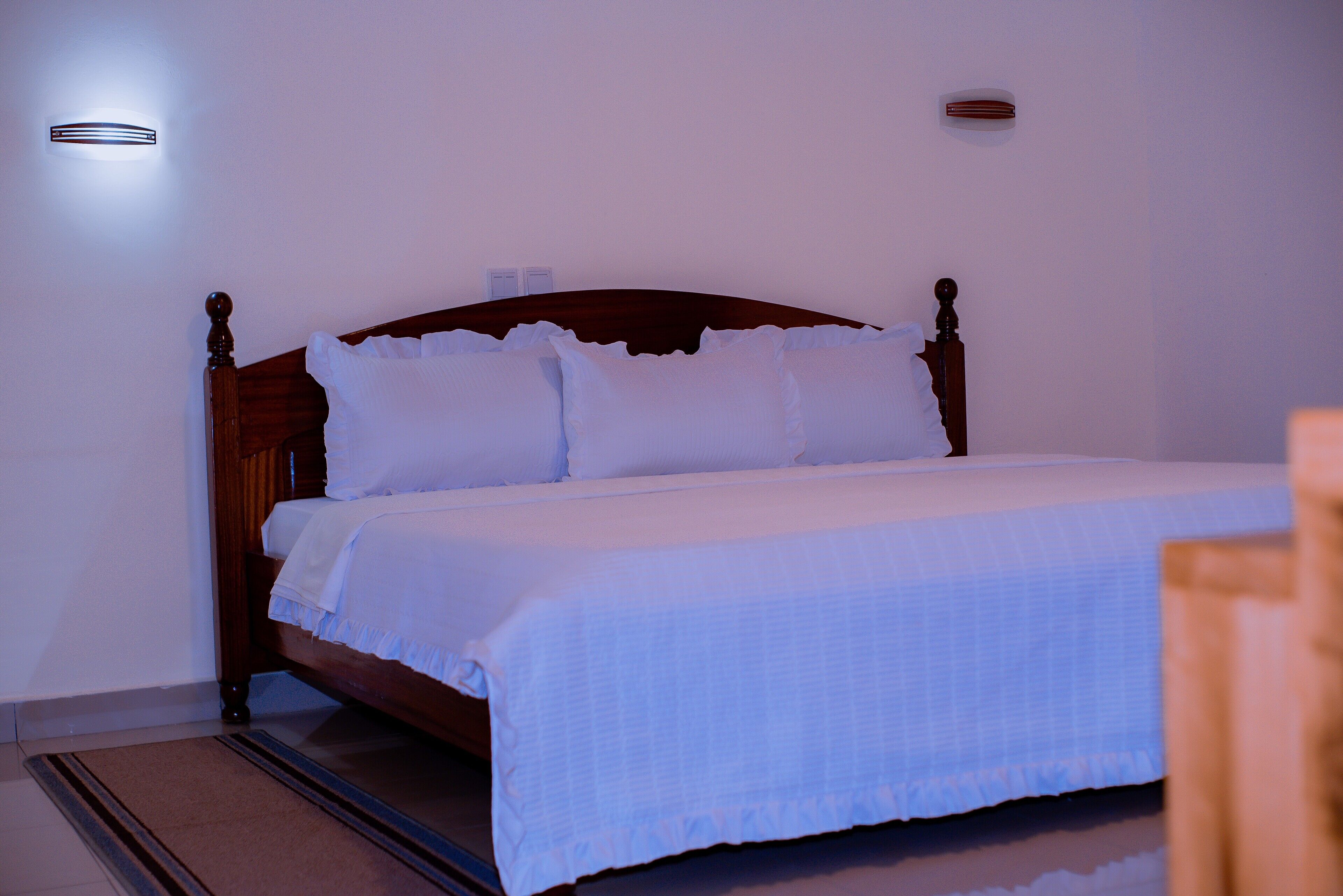 A neatly made bed with white linens and pillows, a wooden headboard, and a small wall-mounted light fixture.