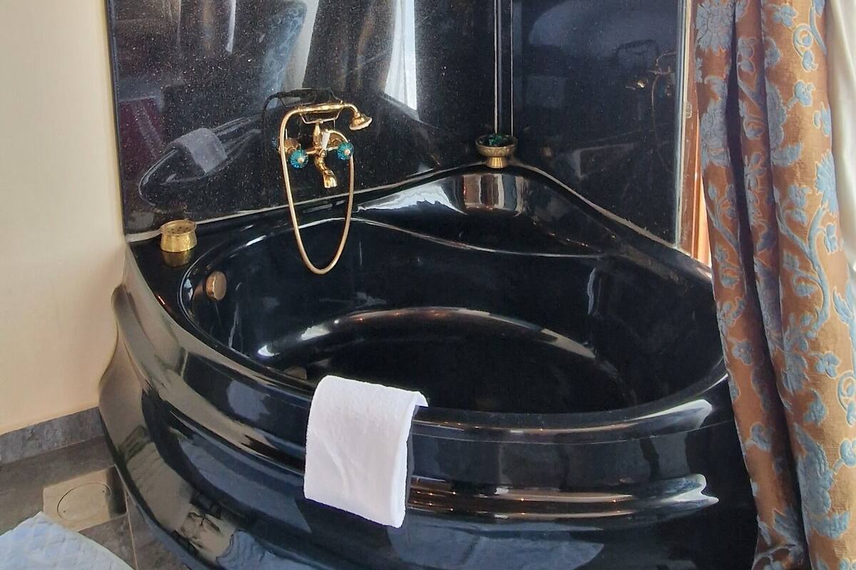 Una bañera de hidromasaje negra con un grifo dorado y una toalla blanca, rodeada de cortinas estampadas y un techo de madera.