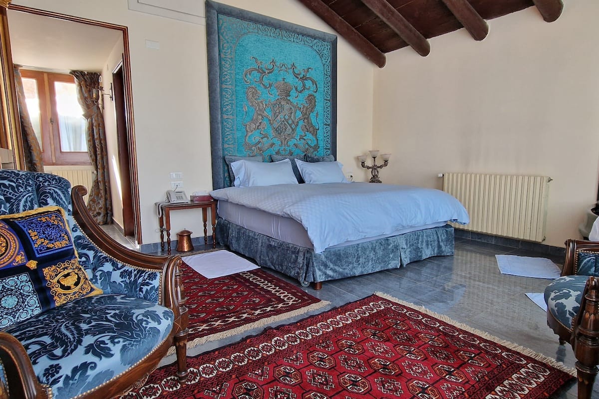 Un dormitorio con una cama, dos sillas, un tapete de diseño rojo y azul, un elemento decorativo grande en la pared y una ventana con cortinas.