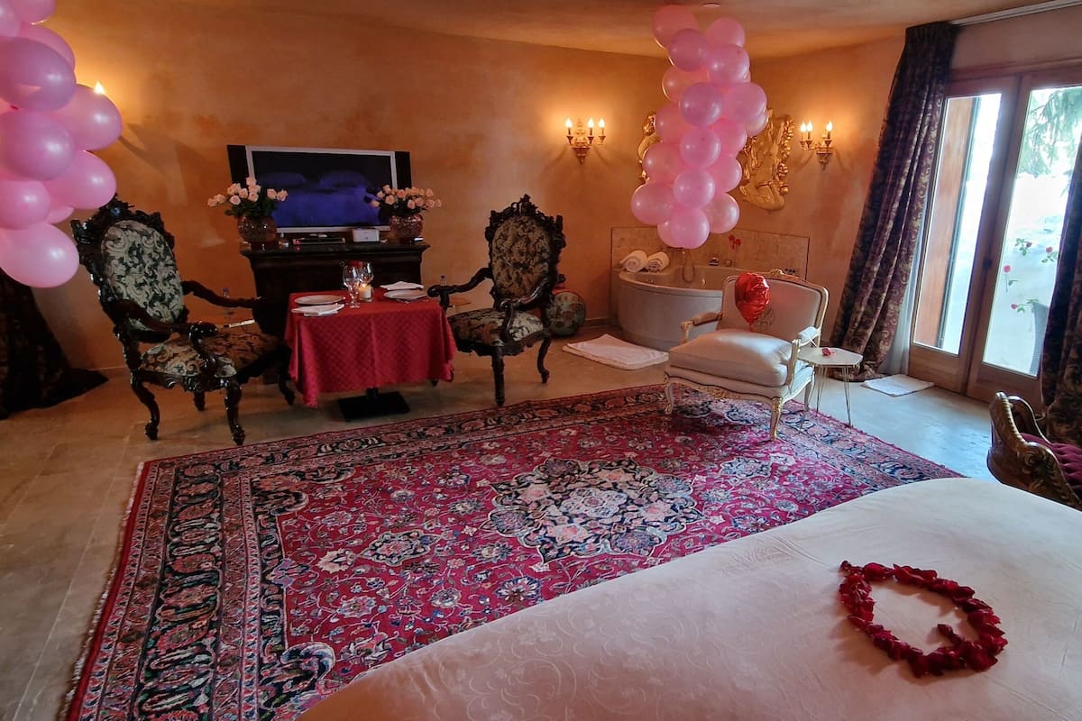 Una habitación de hotel con una cama grande, una alfombra roja, un televisor, una pequeña mesa con mantel rojo y elementos decorativos como globos y un oso de peluche.