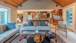 Ein geräumiges Wohnzimmer mit hoher Decke und Holzbalken, einer hellblauen Sofagarnitur, einem Couchtisch und einer Küche im Hintergrund.