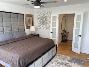 A bedroom with a tufted headboard, a bed with a brown quilt, a bedside lamp, a wall-mounted abstract art piece, and a door leading to another room. - Cozy penthouse Atlanta (Atlanta)