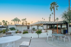 Un espace extérieur aménagé en terrasse, avec des tables et des chaises rondes, un coin salon abrité et des palmiers.