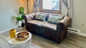 A cozy living room with a brown leather sofa, two green leaf-patterned pillows, a large green plant, a small white round table with a yellow vase and a woven basket, and a floor lamp with three light bulbs.