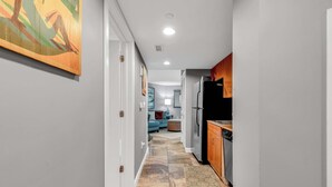 A narrow hallway with a kitchen on one side, a living area with a sofa and a lamp in the background, and a painting on the wall. - Oceanfront Perfect Couples Getaway (Myrtle Beach)