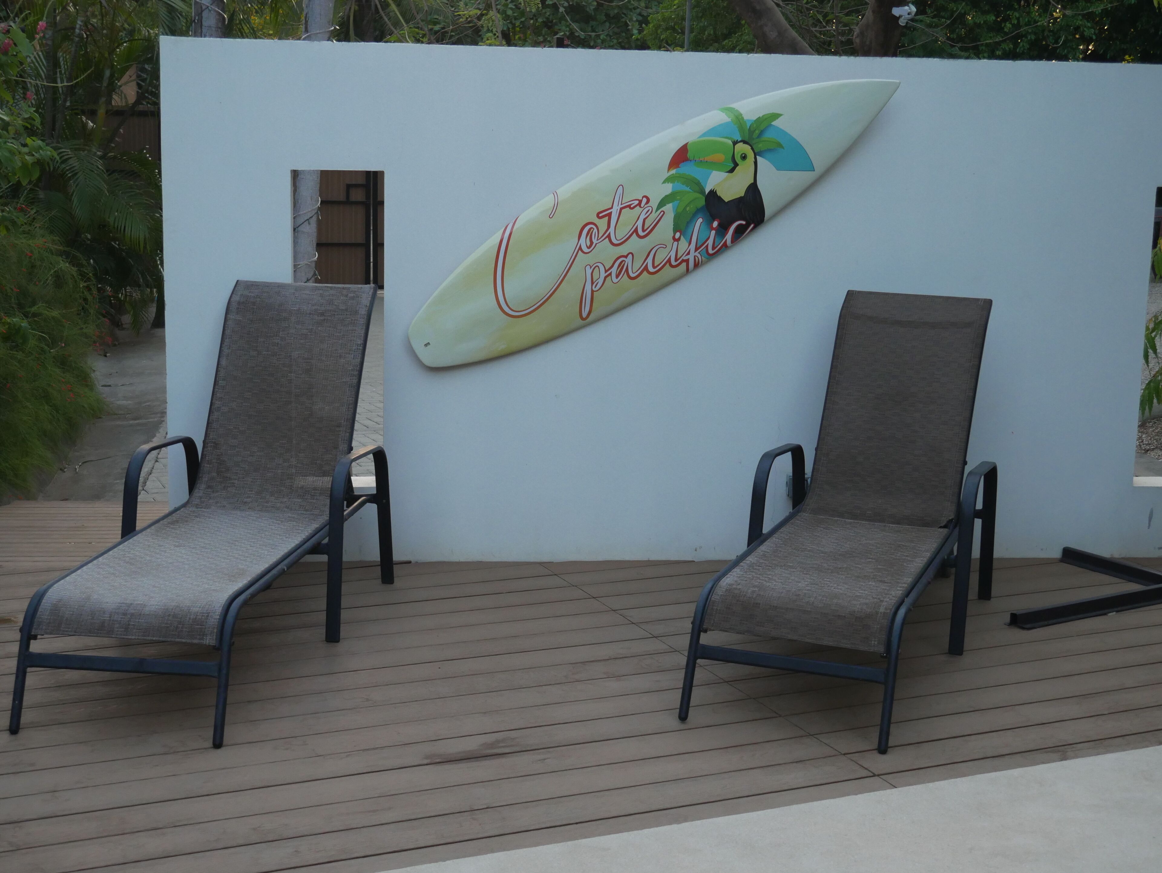 Deux fauteuils de jardin sur une terrasse en bois, avec un mur blanc orné d’un motif représentant une planche de surf et un palmier.