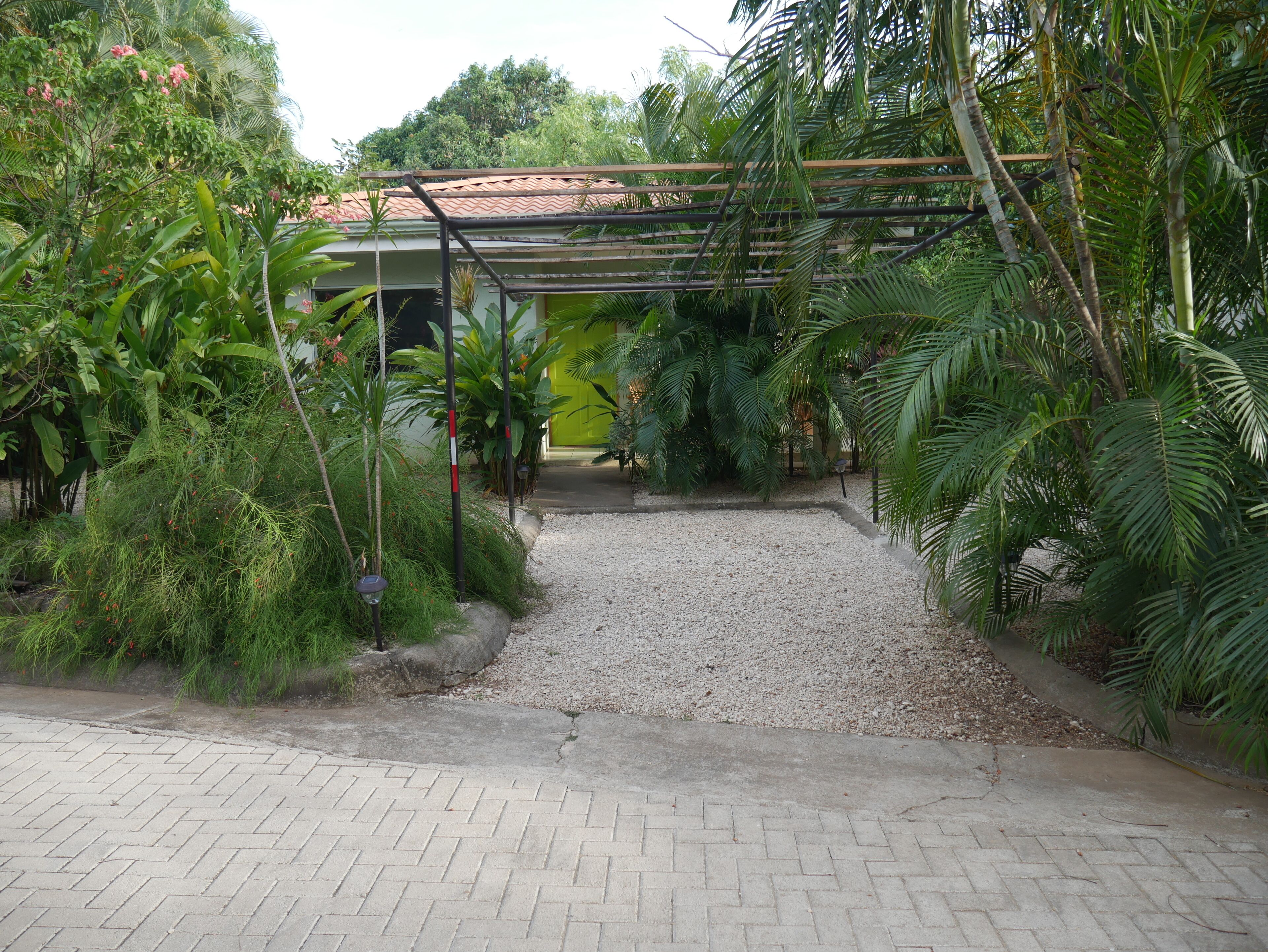 A paved pathway leading to a building with a tiled roof, surrounded by lush greenery and palm trees.