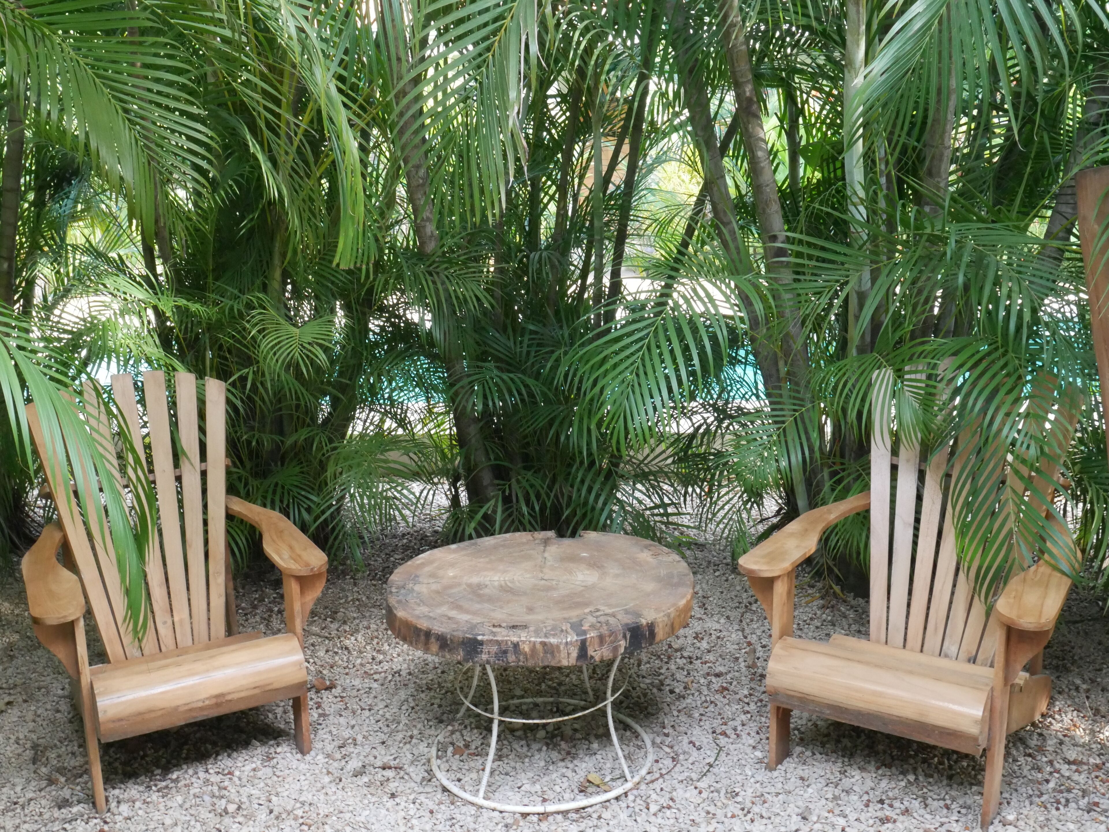 Two wooden Adirondack chairs with a round wooden table in between, set on a gravel surface surrounded by dense green foliage.