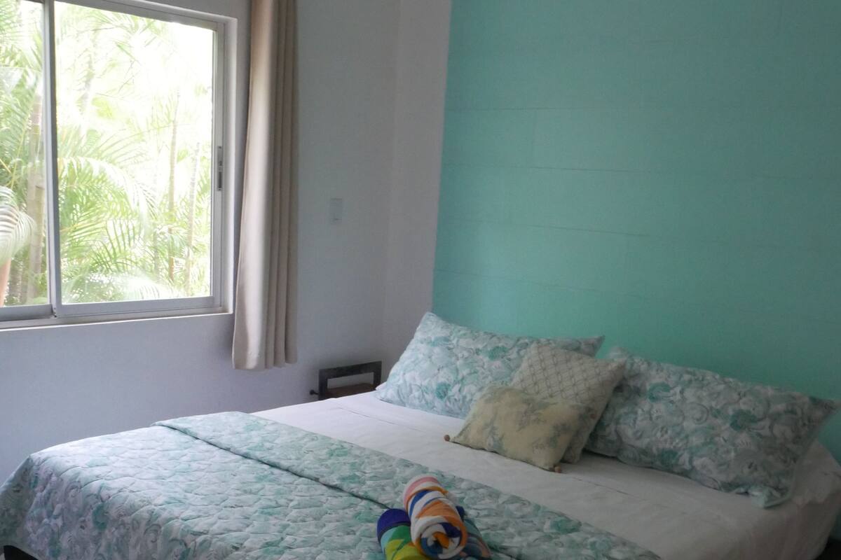 A bedroom with a bed, a window with curtains, and a view of palm trees.