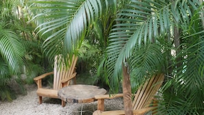 Un espace de détente extérieur en bois, avec deux chaises et une petite table ronde, entouré d’un luxuriant feuillage verdoyant.