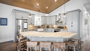 Une cuisine moderne dotée d’un grand îlot central, de tabourets de bar en bois, d’appareils électroménagers en acier inoxydable et de luminaires suspendus.