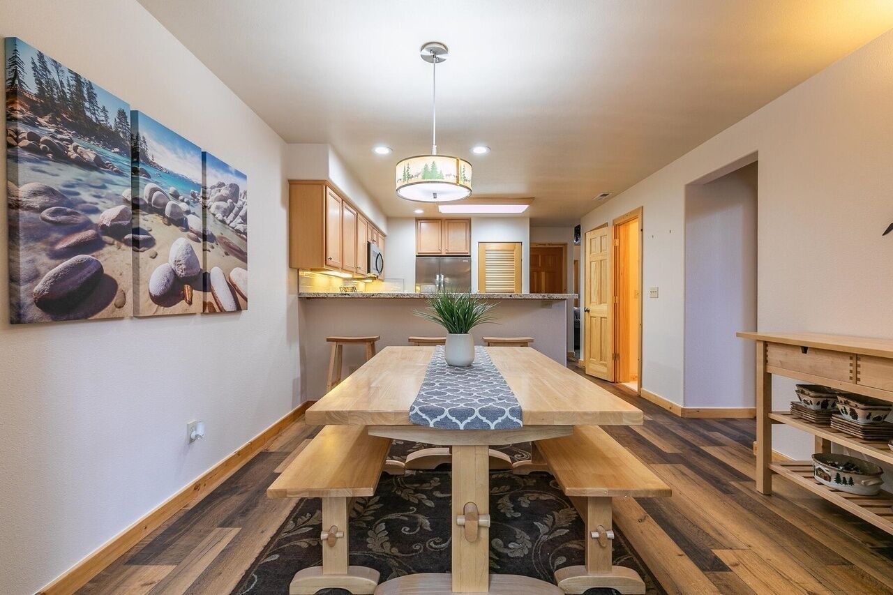 A dining area with a wooden table, benches, and a central plant. There are kitchen cabinets and a refrigerator in the background.