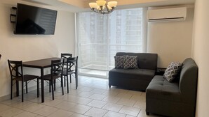 A modern living room with a dark grey sofa, a dining table with four chairs, a flat-screen TV mounted on the wall, and a large window with a view of high-rise buildings. - Apartment in Ecohousing Phase 2 (Tegucigalpa)