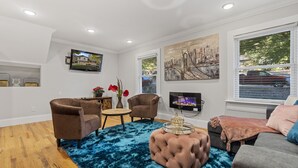 Uma sala de estar moderna com piso de madeira, um tapete felpudo azul, um banquinho estofado, duas poltronas, uma mesa de centro e uma lareira.